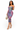 A woman modeling a strapless bodycon midi dress with a purple, pink, and blue tie-dye pattern, paired with gold high-heeled sandals and a woven beige handbag, set against a plain white background.