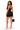 A woman modeling a black strapless bodycon mini dress from the back, holding a black handbag and wearing clear high-heeled sandals against a plain white background.