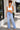 A woman stands on a sidewalk wearing a white ribbed crop top and light-wash blue flare jeans, with her hand in her pocket. The background shows a modern building exterior with a window, potted plants, and a walkway.
