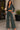 A woman models a dark forest green two-piece lounge set consisting of a cropped button-up top and matching wide-leg pants, standing on a stone patio.
