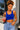 A woman with curly blonde hair wearing a bright royal blue scoop-neck crop top and light-wash blue jeans, holding a pink drink in front of a white brick wall.