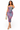 A woman modeling a strapless bodycon midi dress with a purple, pink, and blue tie-dye pattern, holding a beige woven handbag and wearing beige high-heeled sandals against a plain white background.