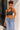 A woman posing outdoors in a dark gray ribbed tank top and light blue high-waisted jeans, standing against a white stucco wall.