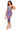 A woman modeling a strapless bodycon midi dress with a watercolor-like abstract print in purple, pink, blue, and green tones, paired with metallic gold high-heeled sandals and a woven beige handbag, against a plain white background.